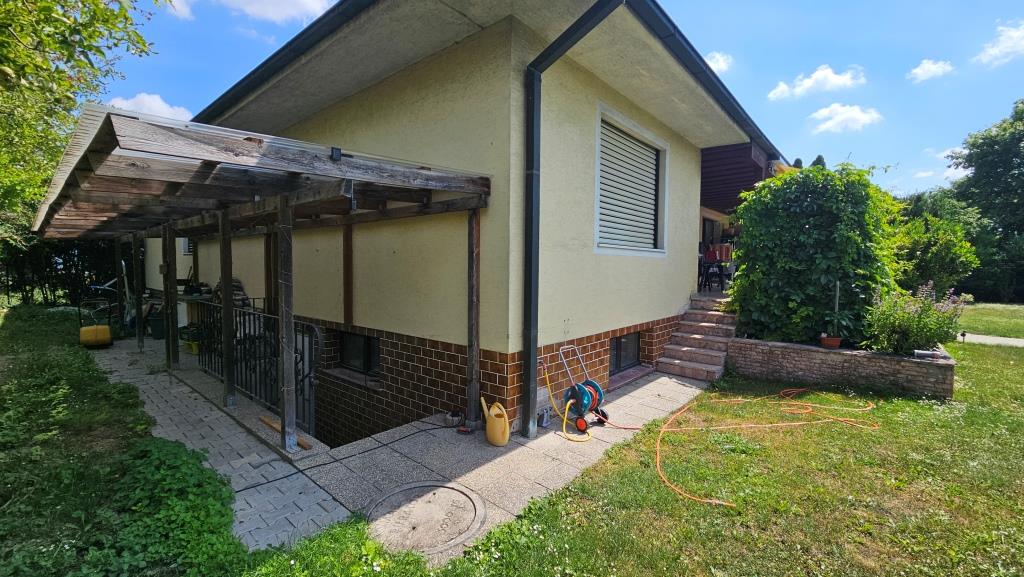 Haus mit gelbem Verputz, Holzbalkon und Garten mit Bewässerungsschlauch