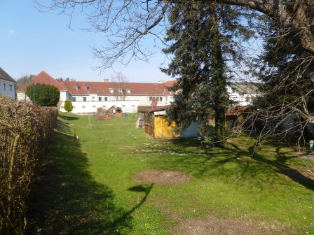 Ländliches Anwesen mit Garten und Holzhaus vor einem langen weißen Gebäude mit rotem Dach