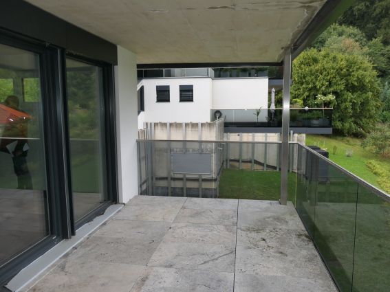 Balkon mit Steinplatten und Glasgeländer vor moderner weißer Villa mit Garten