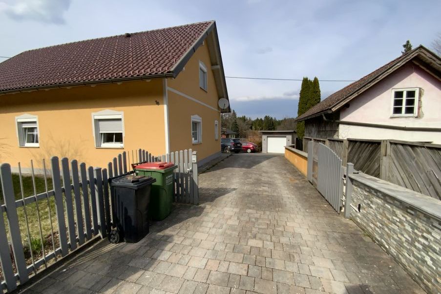 Zwei Mülltonnen vor gelbem Haus mit Einfahrt, Garage und Wald im Hintergrund