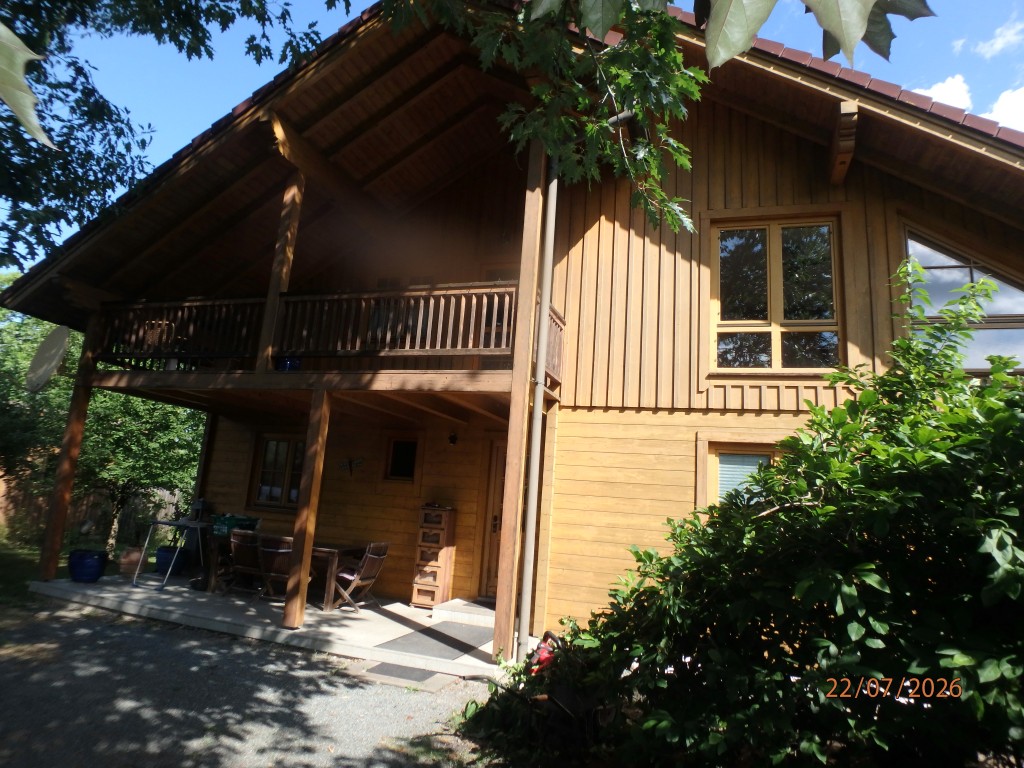 Holzhaus mit Balkon und Terrasse in ländlicher Umgebung, mit Gartenmöbeln und grüner Bepflanzung.