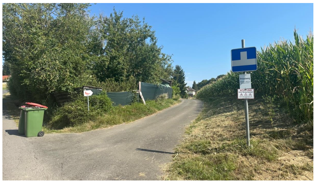 Privatweg-Schild und Mülltonnen an einer asphaltierten Straße mit Maisfeld und Bäumen