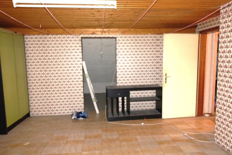 Renovierungsbedarf in einem Raum mit gemusterten Tapeten, schwarzem Regal und Holzboden