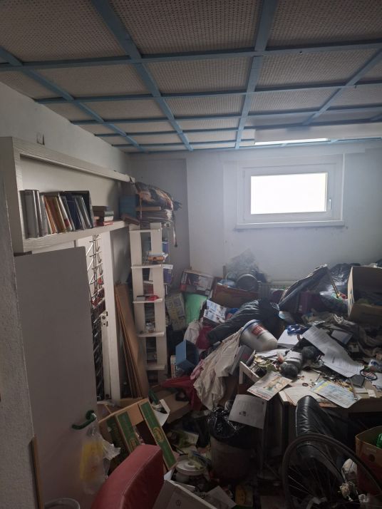 Unordnung in einem Zimmer mit einem Fenster, Bücherregalen und einem Schreibtisch voller Gegenstände.