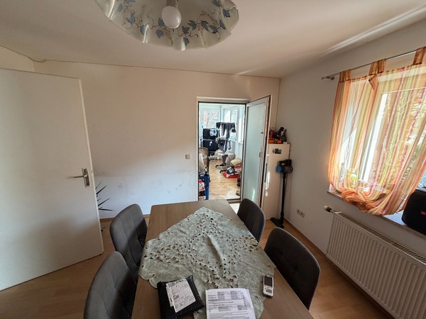 Esszimmer mit Holztisch und vier Stühlen, daneben ein offener Durchgang zu einem weiteren Raum mit Computern.