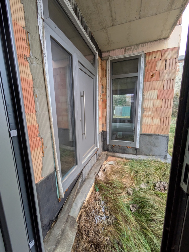 Graue Eingangstür und Fenster an einem Rohbau mit Ziegelmauerwerk und Betonüberdachung.