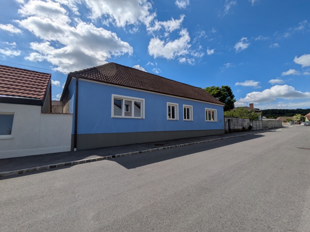 Blaues Haus mit weißen Fenstern und grauer Sockelzone an einer asphaltierten Straße