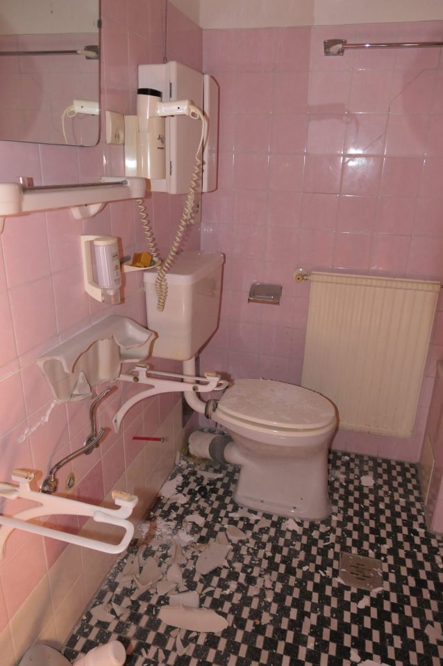 Beschädigtes Badezimmer mit zerbrochenem Waschbecken und Toilette auf kariertem Boden.