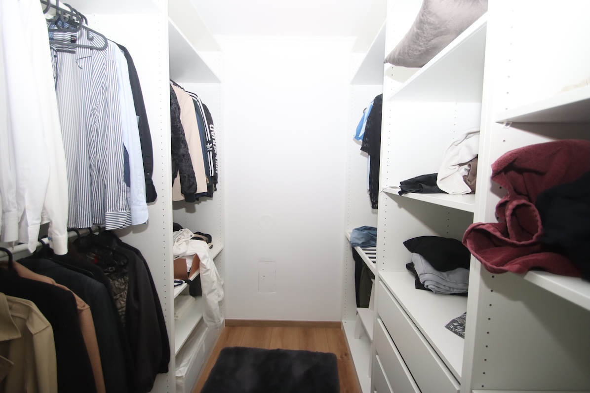 Kleiderschrank mit Kleidung auf Kleiderbügeln und Regalen, ein dunkelgrauer Teppich liegt am Holzboden.