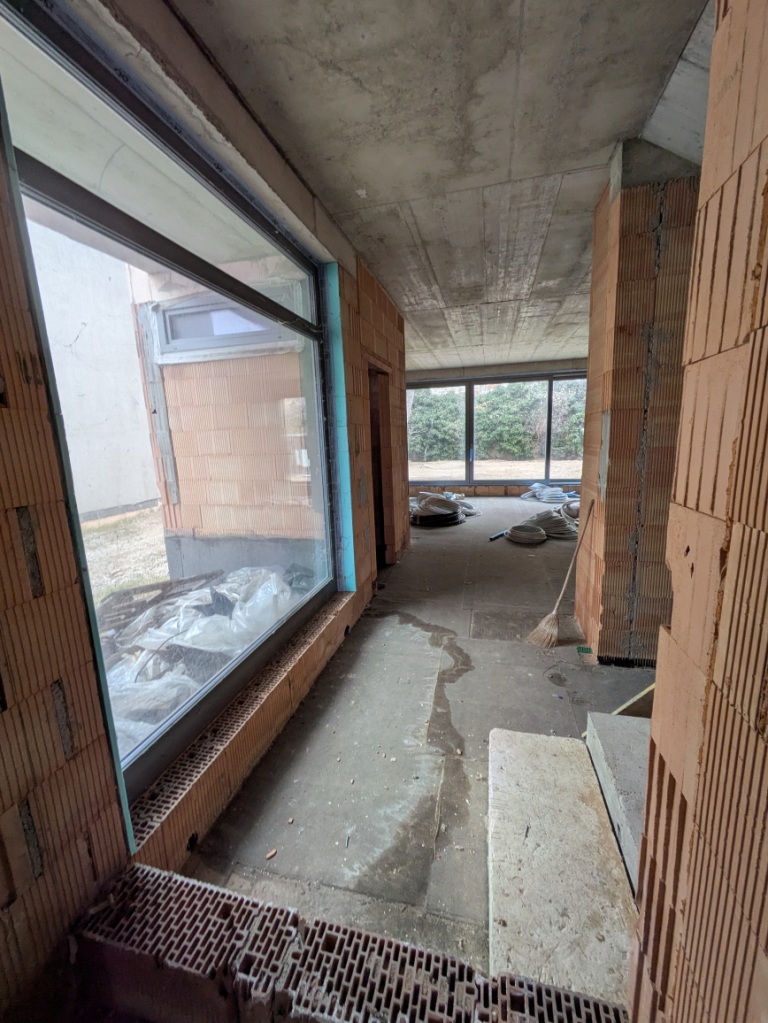 Baustelle mit Rohbauwänden, Beton und großen Fenstern, Besen und Material auf dem Boden