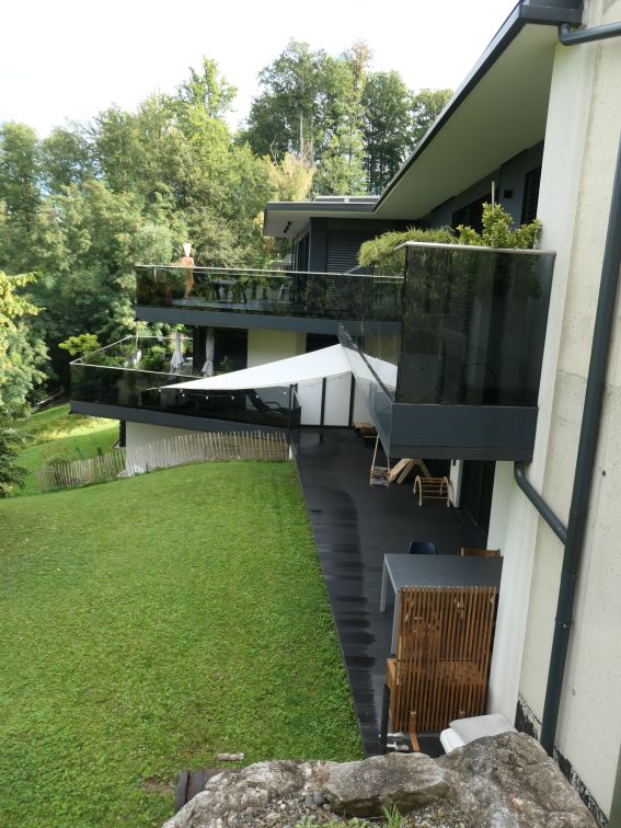 Moderne Villa mit Garten und Terrassen in Hanglage, umgeben von Bäumen.