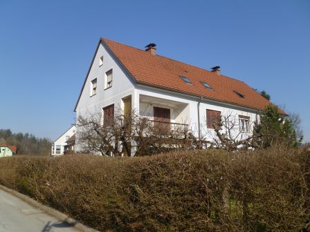 Weißes Haus mit roten Ziegeln und braunen Fensterläden, von einer Hecke verdeckt.