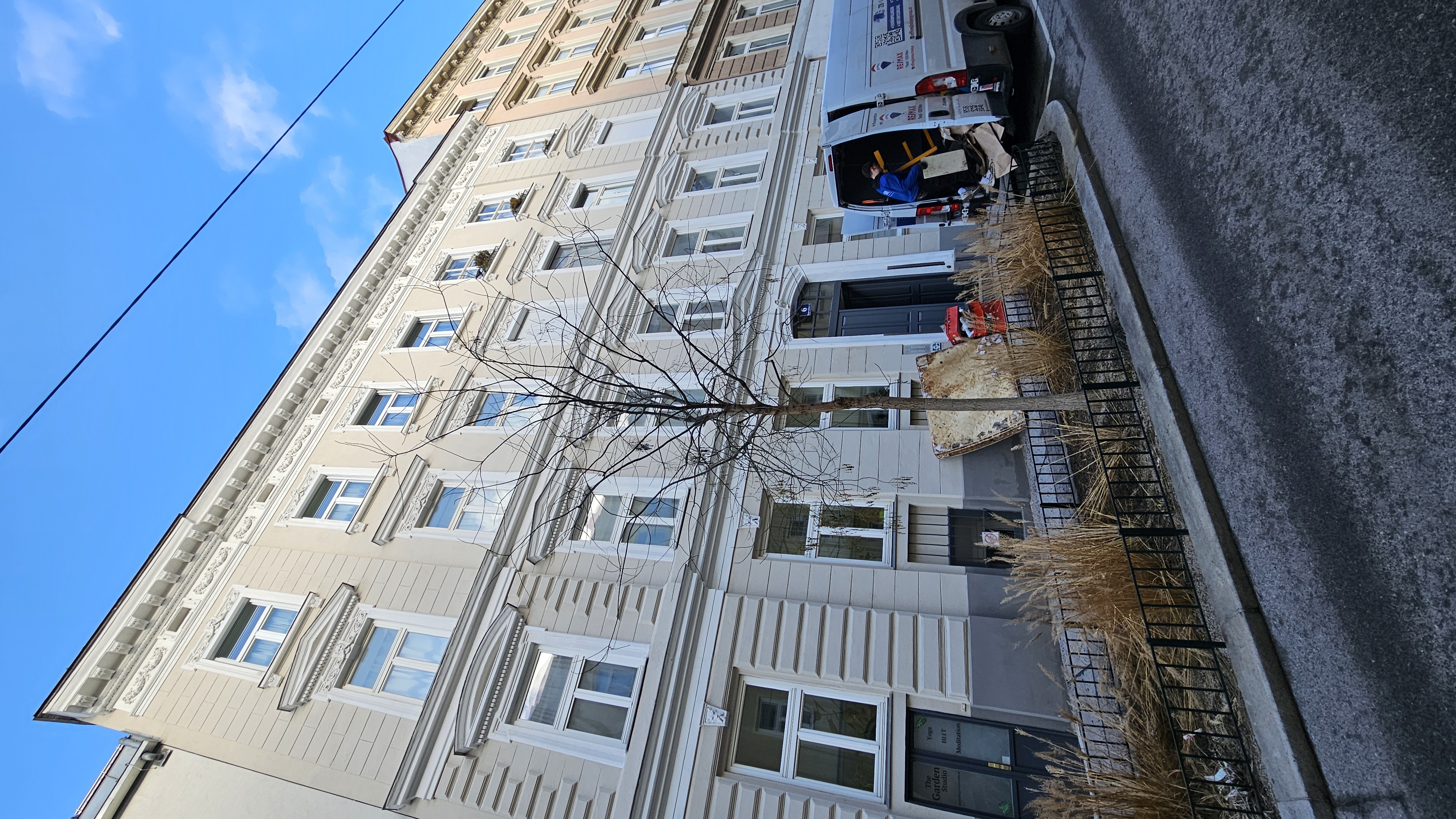 Wiener Wohnhausfassade mit RE/MAX-Lieferwagen und umgestürztem Baum vor einem Geschäft.