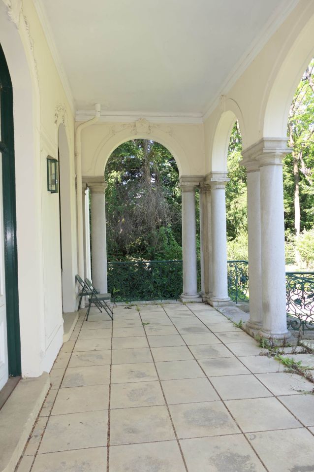Säulengang mit Bögen und zwei Stühlen, Blick auf einen Garten mit Bäumen