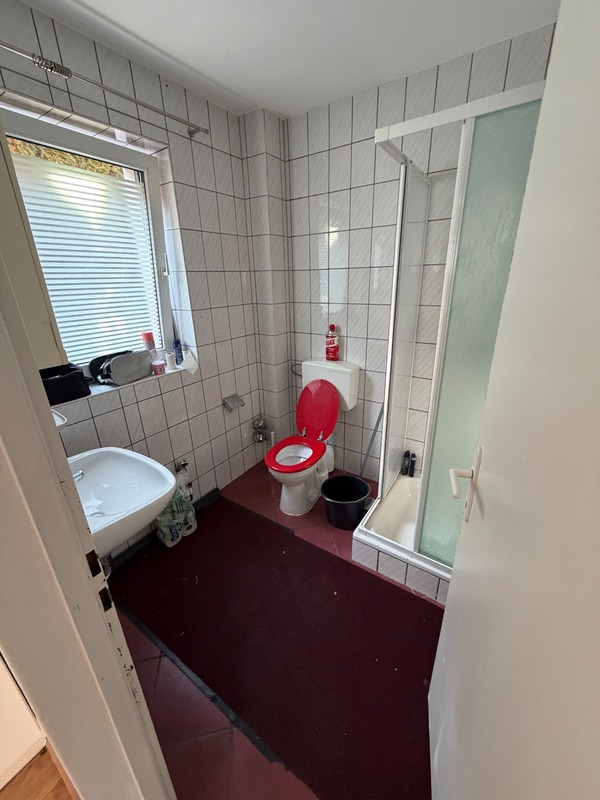 Badezimmer mit Waschbecken, WC mit rotem Deckel, Dusche und Fenster.