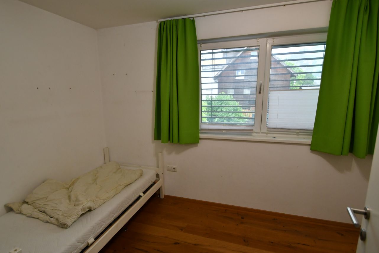 Schlichtes Zimmer mit Bett, grünen Vorhängen und Fenster mit Blick auf ein Holzhaus