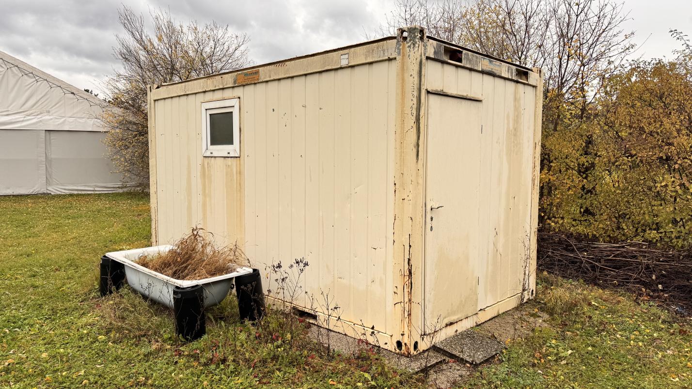 Container mit kleinem Fenster und Tür, davor eine alte Badewanne mit trockenem Gras